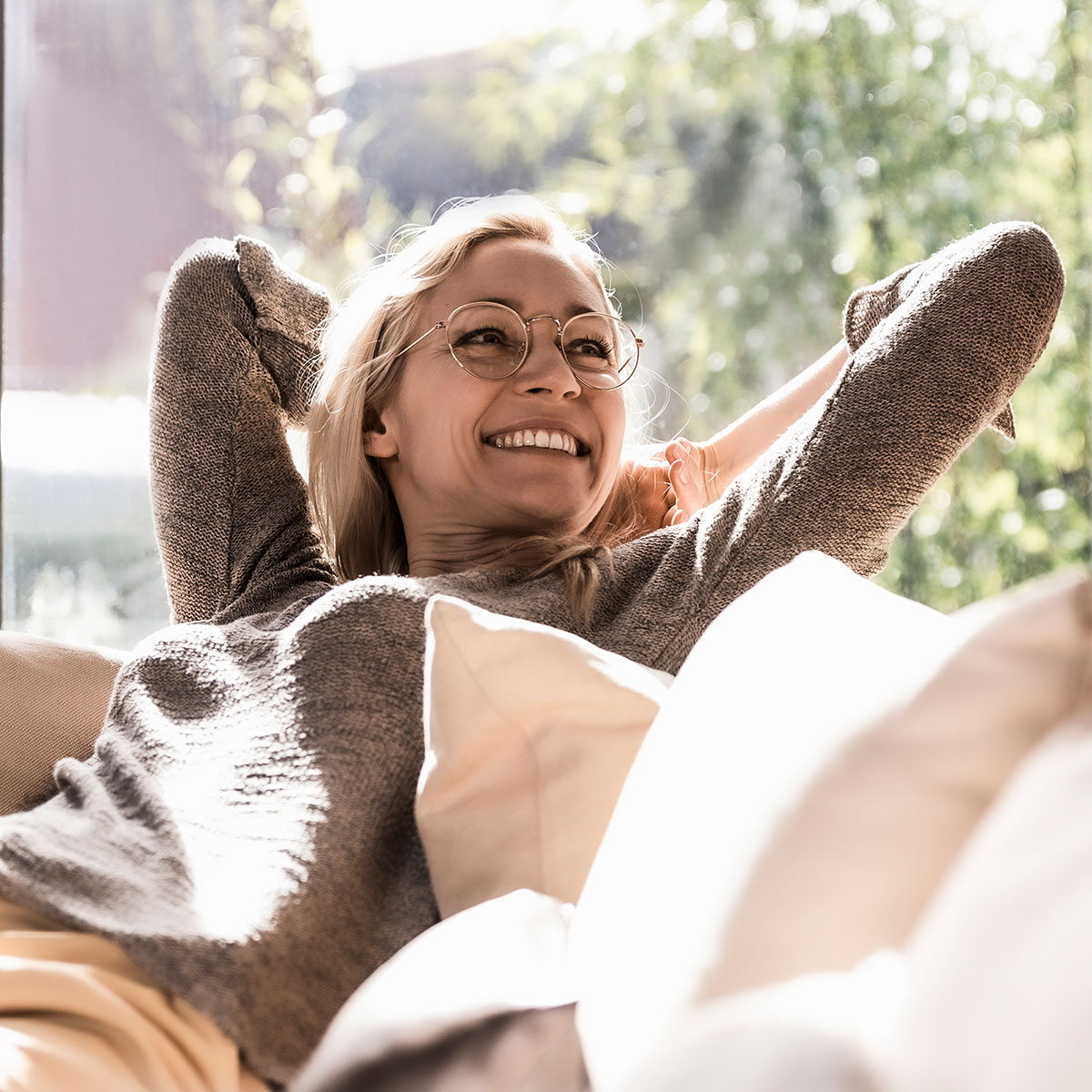 Lächelnde, blonde Frau mit Brille auf dem Sofa mit hinter dem Kopf verschränkten Armen