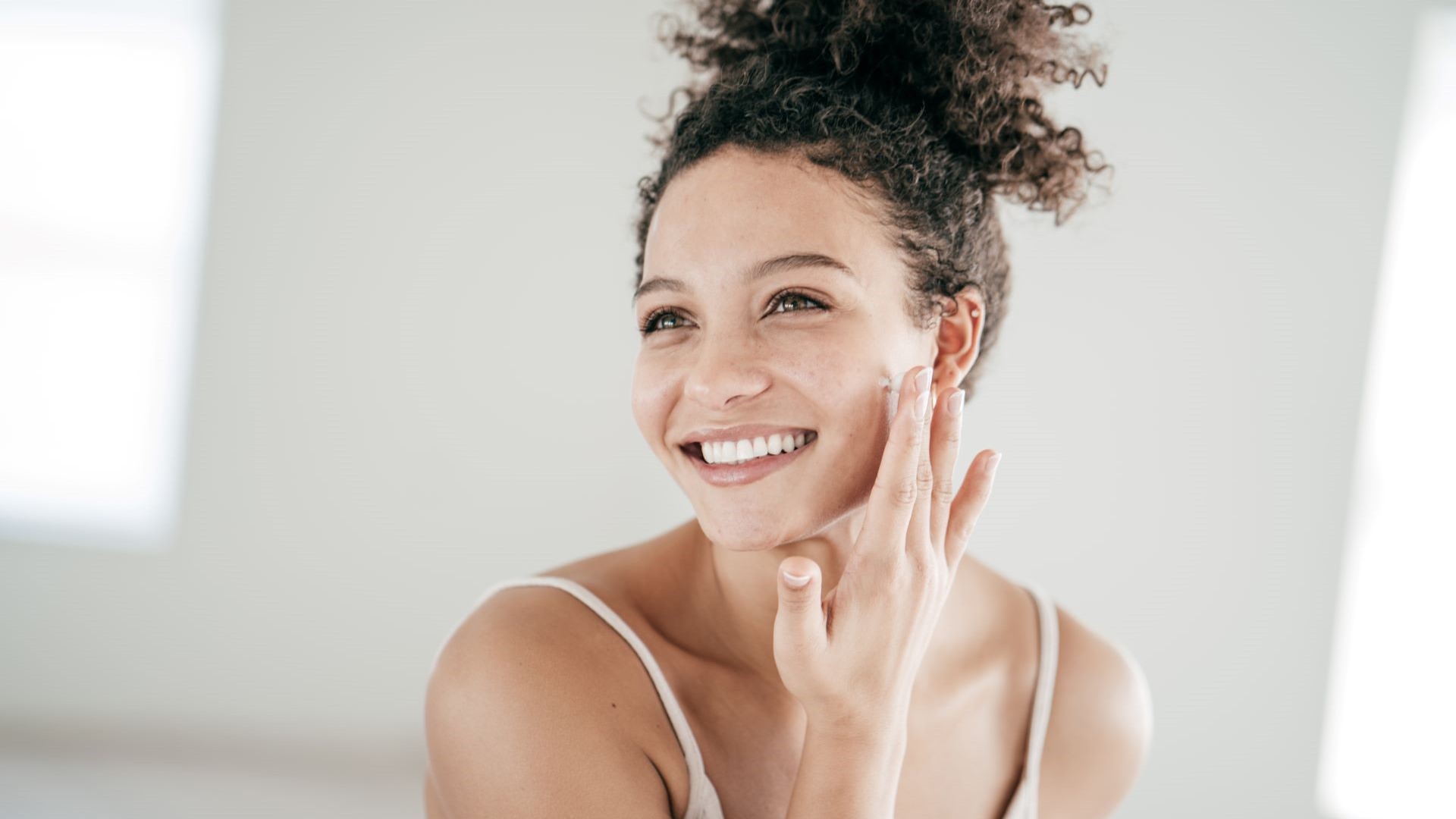 Frau mit braunen Locken lächelt und cremt ihr Gesicht ein